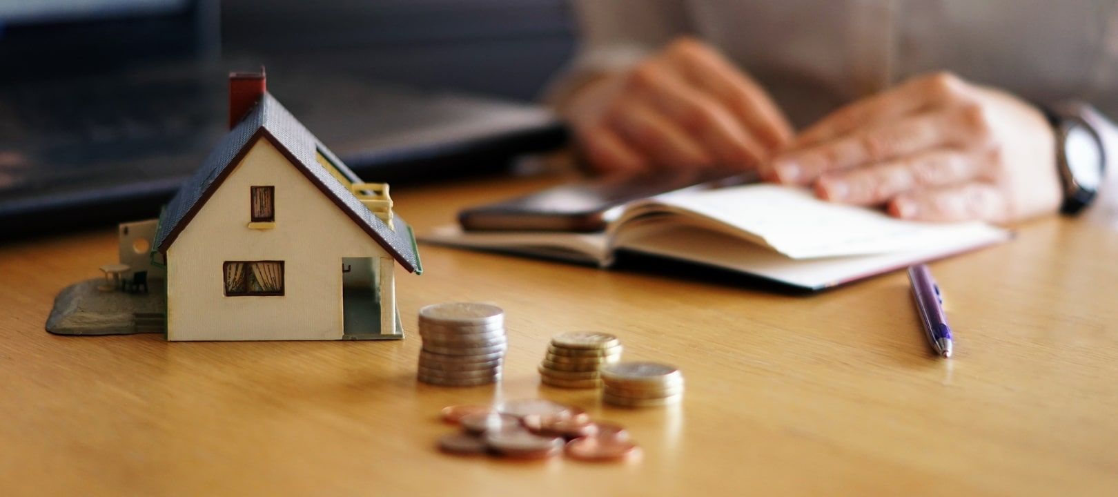 mesa con una maqueta de una casa en la que hay una persona escribiendo en un cuaderno con monedas delante